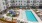  A large swimming pool surrounded by lounge chairs and umbrellas in front of a multi-story apartment building.