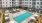  A large swimming pool surrounded by lounge chairs and umbrellas in front of a multi-story apartment building.